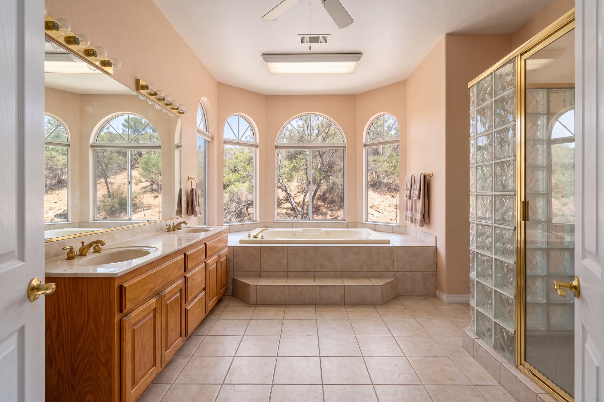 Before: a large 1990s estate primary bathroom with a raised tile platform tub in a bay of five arched windows, a honey oak double vanity under Hollywood strip lights and a glass-block shower