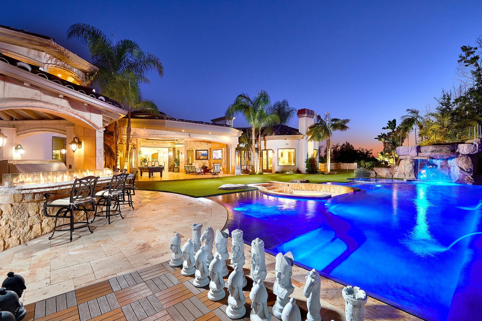 The backyard of a custom San Diego County home at dusk: a pool and spa, a lawn with a giant chess set and the outdoor living room