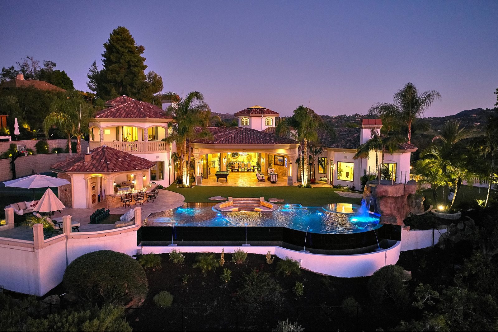 A custom San Diego County home at dusk with a lit pool, a covered loggia and the hills behind
