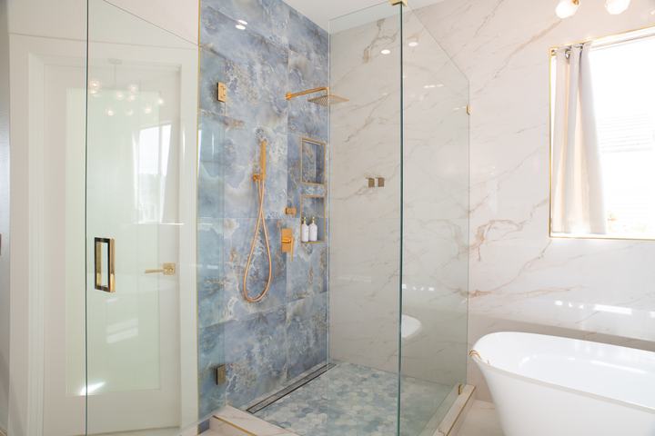 Glass-enclosed shower with a blue feature wall and brass fixtures next to a freestanding tub and marble-look tile walls