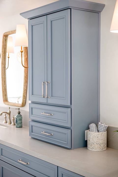 Blue linen tower cabinet with drawers on quartz countertop beside vanity mirror and lighting