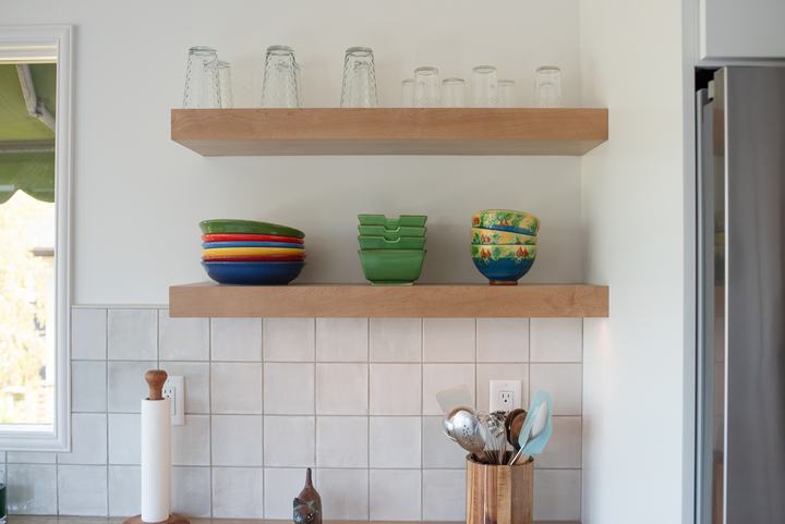 Two maple floating shelves holding glasses and colorful bowls above the backsplash