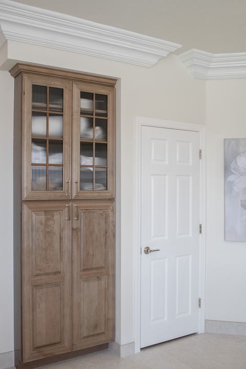 Maple-almond linen cabinet with ribbed-glass doors beside a white interior door and layered crown molding.
