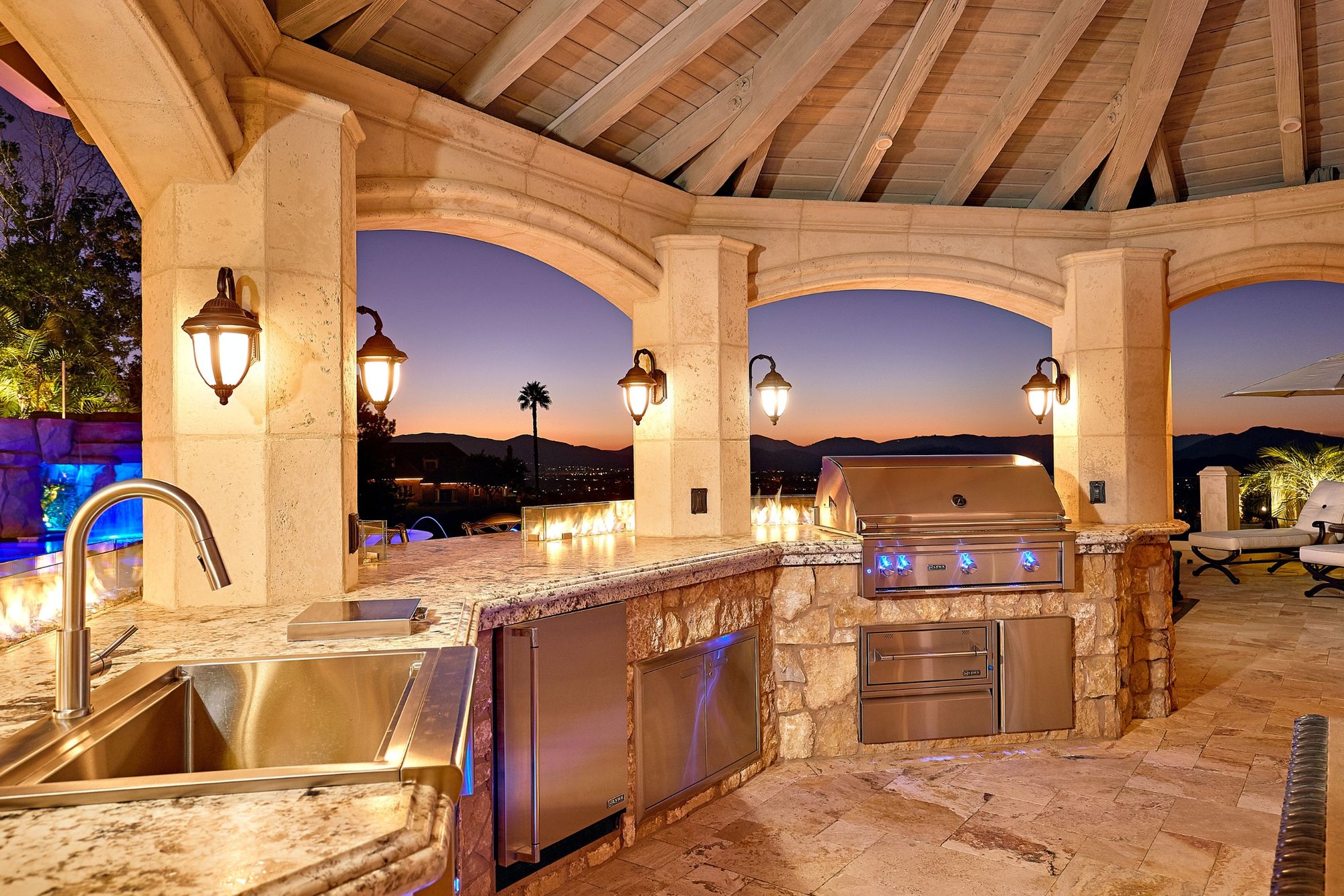 San Diego outdoor kitchen under stone arches at dusk, with a built-in grill, a sink and a view over the pool to the hills
