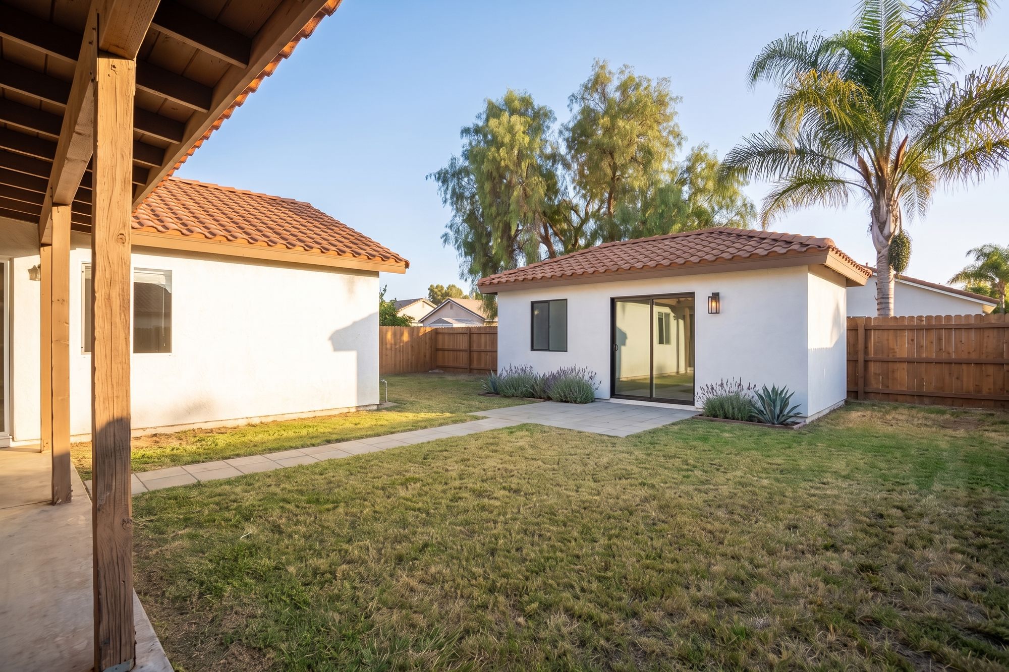 After: the same backyard with a new detached ADU, white stucco and a matching clay tile roof, a black-framed sliding glass door, a paver patio and a path across the lawn