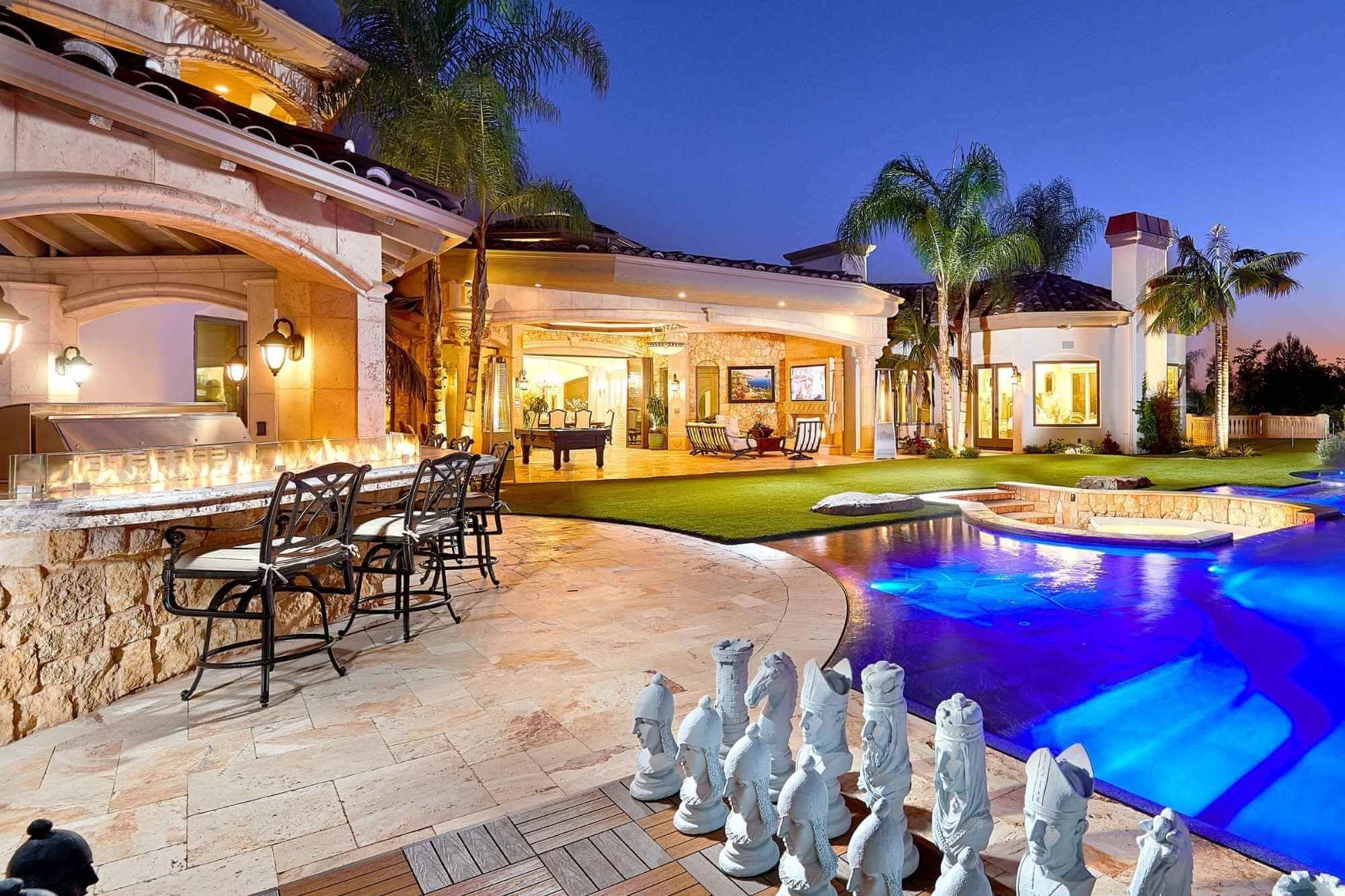 After: the same backyard at dusk with a new pool and spa, a lawn with a giant chess set, a stone outdoor kitchen under arches and a covered outdoor living room opened to the yard