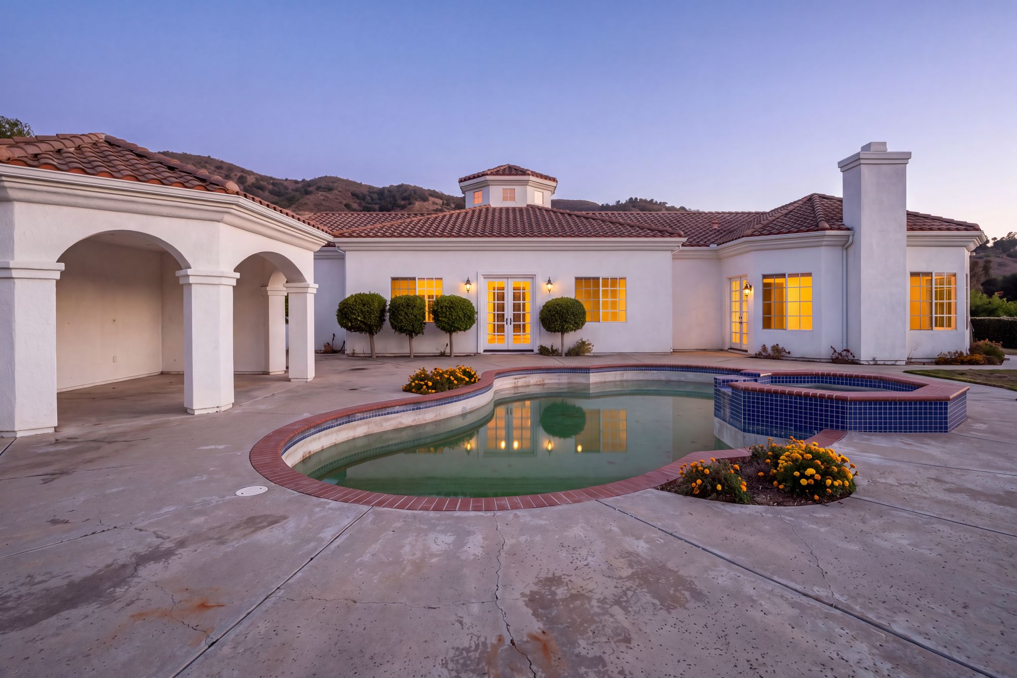 Before: the same San Diego backyard at dusk as it was, a worn, unlit free-form pool with red brick coping and a blue-tile spa on cracked concrete, a plain white stucco cabana, and the original house wall with French doors where the outdoor living room now stands