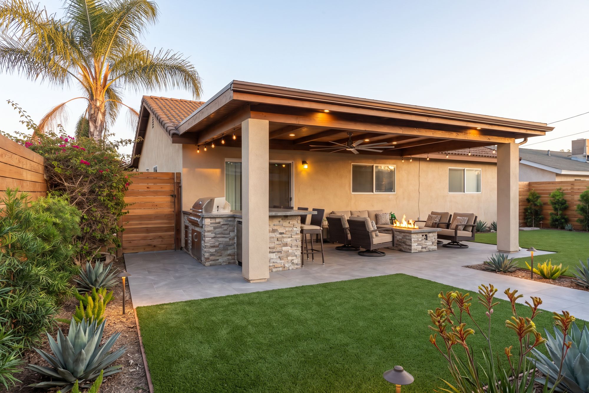 After: the same backyard with a covered California room on a beamed ceiling, a stone outdoor kitchen, a lounge around a gas fire pit, porcelain pavers, turf and a new cedar fence