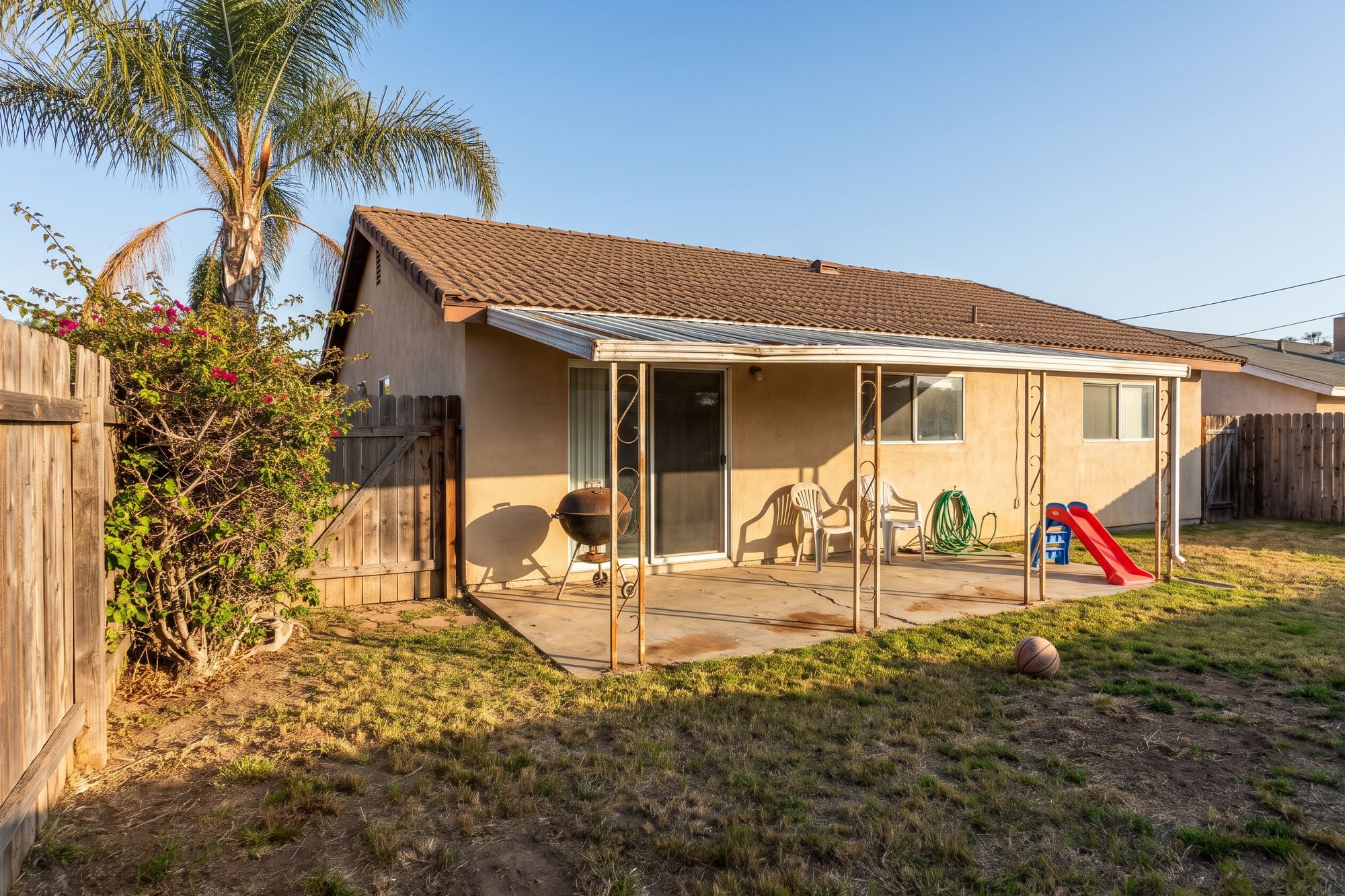 Before: a lived-in San Diego tract-home backyard, a sagging aluminum patio cover over cracked concrete, a patchy lawn, a kettle grill, plastic chairs and a kid's slide